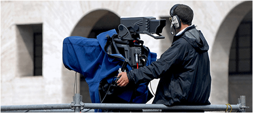A camera operator focuses a large professional video camera while preparing for a broadcast, set against a historic architectural backdrop.